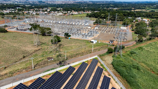 A high angle aerial view showing photovoltaic solar panels in the foreground with a large electricity substation and power grid in the background in Atalaia, Goias, Brazil.