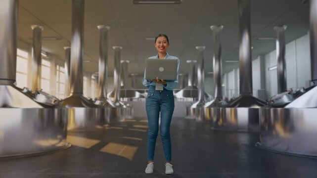 Full Body Of Asian Female With Her Laptop at Stainless Steel Brewing Vats in Beer Plant, She Is Looking At The Camera With A Smile