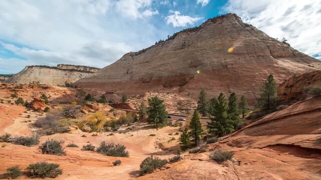 A wide shot captures the scenery of Zion National Park in Utah. Towering, ancient escarpments of red and cream-colored Navajo sandstone dominate the frame under a bright blue sky with scattered white 