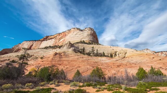 An establishing shot of massive, layered red and white sandstone formations under a bright blue sky with dynamic white clouds in Zion National Park, Utah, USA.