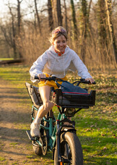 Fototapeta premium Happy laughing young woman riding fast on an electric cargo bike along path. Active urban lifestyle, eco-friendly transportation and sustainable mobility concept.