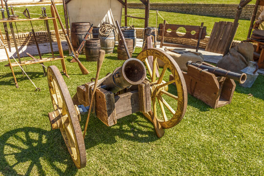 Wide shot of vintage field cannons with wooden wheels, alongside historical barrels and a pillory at a medieval battle reenactment festival.