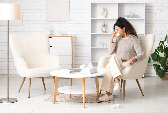 Young African-American woman sneezing in handkerchief and sitting on armchair at home. Allergy concept