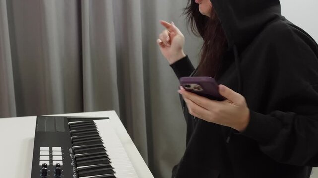 Woman in hoodie composing music. Casual woman using phone while creating music. Person dressed comfortably in hoodie engaged in music sampling and arrangement at small studio