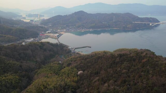 Aerial tilt up from bird's eye view of Hirakiyama mountain top to Omishima Bridge in morning haze. Panoramic vista over Seto Inland Sea, Japan.
