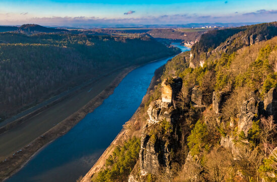 Downstream view along the Elbe River toward the Wartturm, the collapsed sandstone tower, in a picturesque panoramic winter scene with blue sky and golden sunrise light above the Bastei cliffs.