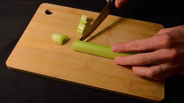 a person&rsquo;s hands using a professional knife to slice a fresh, green celery stalk into uniform pieces on a bamboo cutting board,
