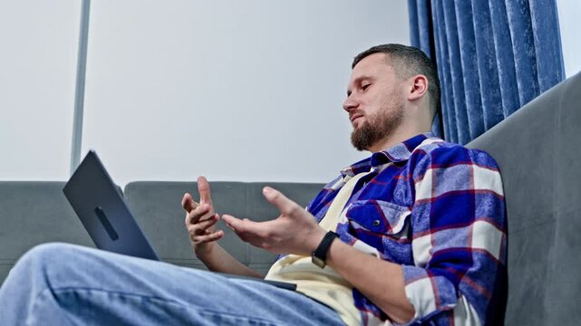 A man is sitting on a couch and talking on a computer. He gestures with his hands while looking at the screen. The room has blue curtains letting in light.