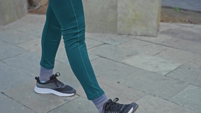 Woman walking forward in teal leggings with black sneaker stepping on stone street pavement; urban motion energy.