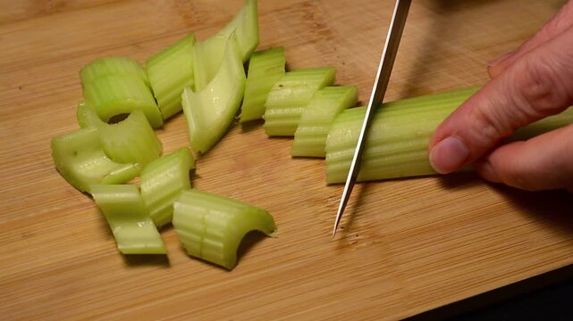 a person&rsquo;s hands using a professional knife to slice a fresh, green celery stalk into uniform pieces on a bamboo cutting board,