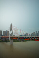 Fototapeta premium Red bridge spans wide river under cloudy sky. Modern structure rises from calm, muddy waters. City skyline fades softly in hazy background. Steel cables stretch from tall central pylon