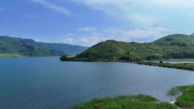 Cinematic drone flight over the calm surface of the Bukhtarma Reservoir. The footage features lush green grassy shores on both sides and low rolling green mountains ahead. A clear summer day