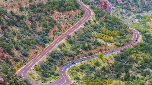 Aerial view footage captures the dramatic switchbacks of the Zion-Mt. Carmel Highway (Route 9) cutting through the rugged, forested landscape of Zion National Park on a bright, sunny day.