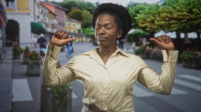 Woman with thumbs pointing to chest on a street crosswalk, wearing beige shirt near outdoor cafe tables and planters in an urban plaza; confidence empowerment.