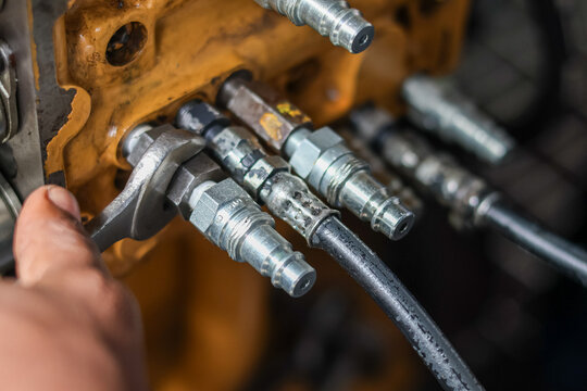 Close-up of a mechanic hand using a wrench to tighten hydraulic hose fittings on a yellow industrial engine block.