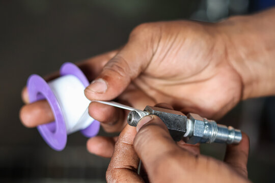 Close-up of mechanic hands wrapping white teflon seal tape on a hydraulic fitting connector thread in a workshop to prevent leaks.