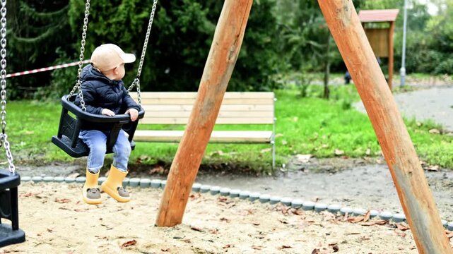A boy of 2 years old swings on a swing in a city park. Toddler swings in the park on a playground. The child plays outdoors and has fun. The boy is dressed in a demi-season jacket and cap.
