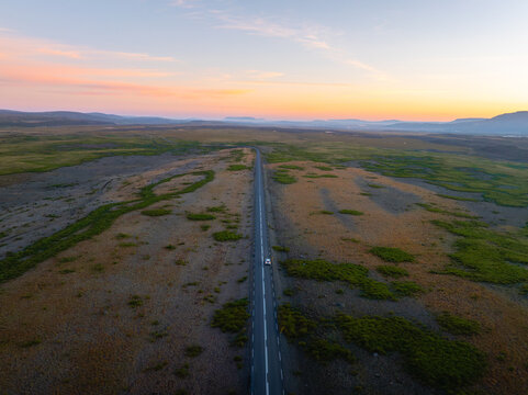 Aerial view of a single road cutting through a vast landscape, the sky painted with hues of orange and blue, Reykholt, Iceland.