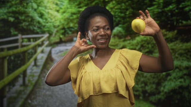 Woman holding lemon with raised right hand and call me gesture at ear, wearing yellow dress on a forest path with wooden railing, smiling; playful joy.
