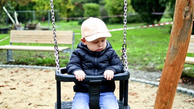 A boy of 2 years old swings on a swing in a city park. Toddler swings in the park on a playground. The child plays outdoors and has fun. The boy is dressed in a demi-season jacket and cap.