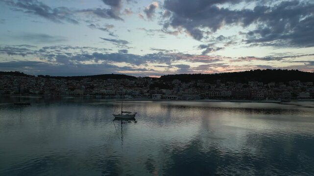 A solitary sailboat drifts on calm waters as dusk settles over a coastal town, with a soft, cloudy sky reflecting in the sea.