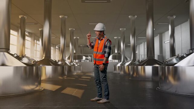 Full Body Side View Of Angry Asian Male Engineer With Safety Helmet Shouting At Someone While Standing at Stainless Steel Brewing Vats in Beer Manufacturing Plant