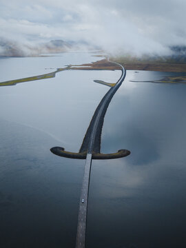 Aerial view of a captivating sword bridge stretches across the tranquil water, connecting landmasses under a blanket of ethereal mist, Snaefellsnes, Eyja- og Miklaholtshreppur, Iceland.