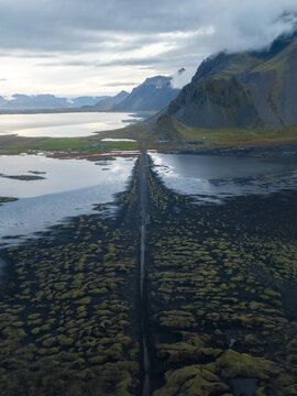 Aerial view of the stark, black sands meeting the tranquil waters near Vestrahorn Mountain, under a brooding sky, Hofn, Iceland.