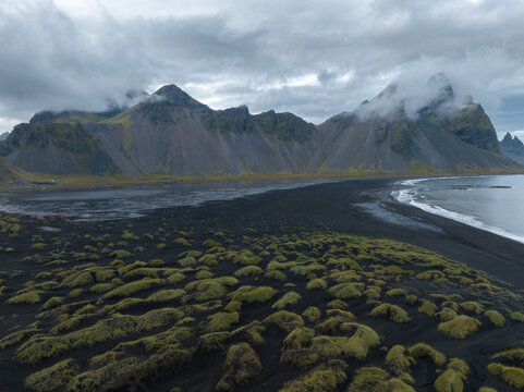 Aerial view of dark sands meeting the sea beneath the towering Vestrahorn mountain range under a brooding sky, Hofn, Iceland.