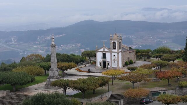 Drone aerial view of white baroque chapel with bell tower and stone column on forested hill