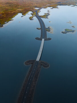 Aerial view of a striking road cuts through the tranquil waters, a ribbon of grey against the deep blue, connecting patches of land under the expansive sky, Westfjords, Iceland.