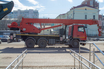 Fototapeta premium Red mobile crane parked on city street construction site. Heavy machinery for lifting load and building architecture. Industrial vehicle for urban development and infrastructure project.