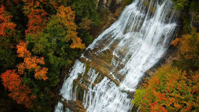 Aerial view of cascading Herisson Waterfalls amidst the vibrant autumn foliage, a tapestry of orange and green hues, Le Frasnois, Bourgogne-Franche-Comte, France.