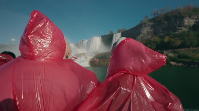 Tourists in red rain ponchos watching Niagara Falls waterfall during boat tour with rainbow in mist