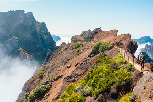 A lone hiker follows a narrow, railed path along a dramatic volcanic ridgeline in Madeira, where steep cliffs drop away into a soft valley of clouds.
