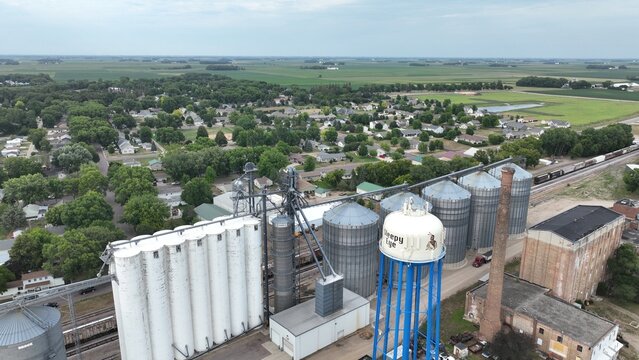 Aerial view of grain elevators and a water tower rise amidst a landscape of green fields and clustered houses, Waseca, Minnesota, United States.