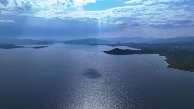 Dramatic aerial drone view of the vast blue Bukhtarma Reservoir. Spectacular sunbeams break through the clouds, illuminating the rolling hilly shores of East Kazakhstan. 