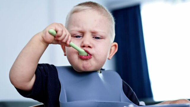 A boy sits at a table and eats food from a bowl using a spoon. He wears a bib and shows an expression while enjoying his meal. Sunlight comes through the window.