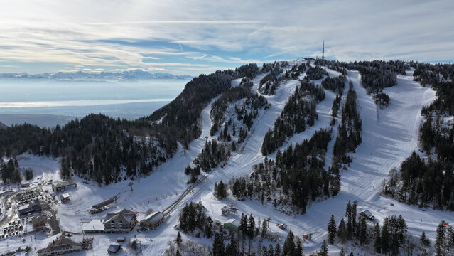 Aerial view of snow-covered slopes descend from a peak crowned with a telecommunications tower, contrasting with the dark evergreens and distant mountains, Gex, Auvergne-Rhone-Alpes, France.