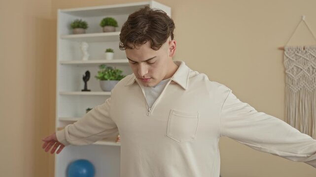 Man young hispanic standing with arms outstretched and hands visible beside an exercise ball, shelf and macrame wall hanging in a studio; calm.