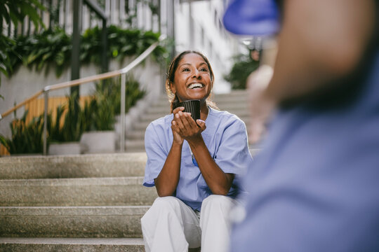 Happy female nurse holding coffee mug sitting on stairs while enjoying leisure time with coworker in hospital building