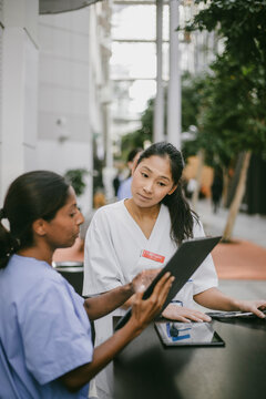 Female healthcare worker looking at documents held by coworker while discussing medical papers in hospital lobby