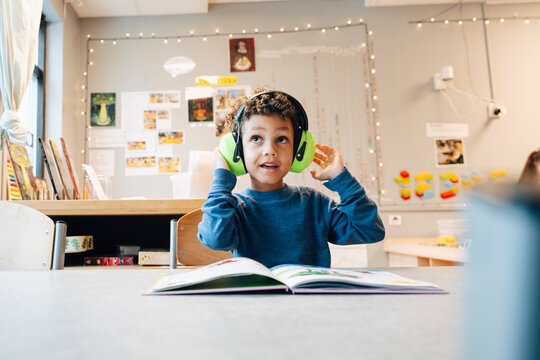 Boy looking away wearing ear muff while sitting at table with book in classroom