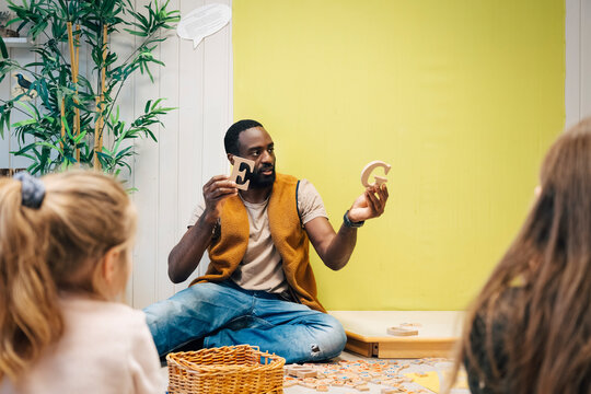 Male teacher holding wooden letters E and G while interacting with children at preschool