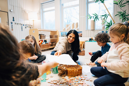 Smiling female teacher passing sheet of paper to girl during fun activity in preschool classroom