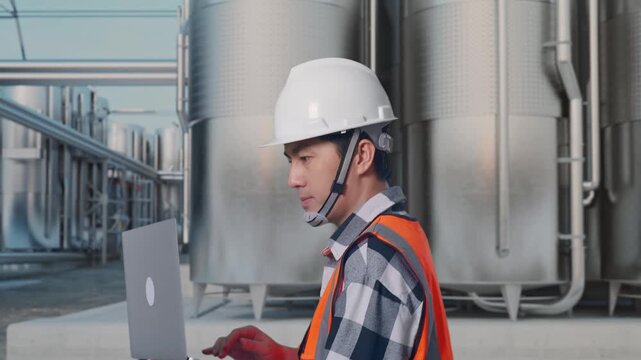 Side View of Asian Male Engineer Use Computer While Walking at Industrial Chemical Plant with Row of Stainless Steel Storage Silos