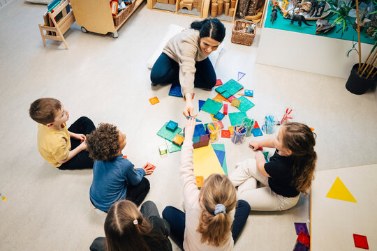 High angle view of female teacher passing geometric shape to girl during fun activity in classroom