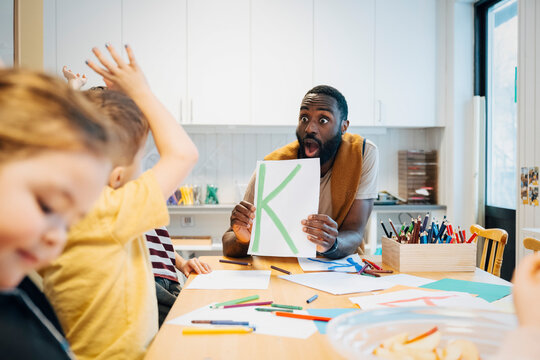 Mature male teacher making surprised face holding letter K during group reading session with kindergarten students