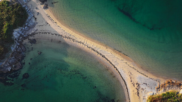 Aerial view of the sandy isthmus connecting rocky outcrops amidst the shimmering, emerald and turquoise waters, Carantec, Brittany, France.