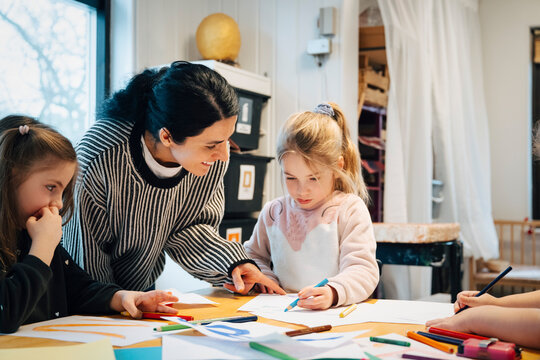 Smiling female teacher assisting kindergarten students during creative lesson in classroom
